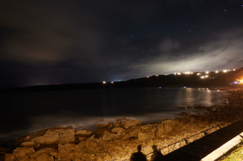 Nighttime photographers The landscape photograph shows nighttime photographers at the coast of Sennen Cove in Cornwall. Taken at night during the autumn season, the image captures the rocky shoreline under a dimly lit sky, with a faint glow from the lights of houses spread along the hillside in the distance. The shadows of the photographers can be seen in the foreground, cast by artificial lighting along the coastal path. The sky above Sennen Cove is partially cloudy, with stars visible in the clearer areas. The photograph highlights the tranquil atmosphere of the Cornish coast at night, with gentle waves creating a misty effect on the water’s surface near the rocks.